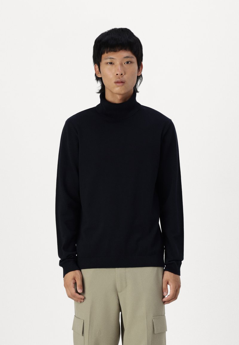 Jersey de cuello alto negro con mangas largas y puños acanalados. Hecho de una tela suave, diseñado para ajustarse cómodamente, usado con pantalones de color claro.