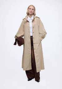 Γυναίκα που φορεί ένα μπεζ μακρύ παλτό πάνω από λευκό πουκάμισο και καφέ φαρδιά παντελόνια, κρατώντας ένα διπλωμένο καφέ μπουφάν στο αριστερό της χέρι.