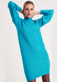 Türkises Strickkleid mit hohem Kragen, Puffärmeln und zwei dekorativen Knöpfen auf der Schulter. Weiche Textur und knielanges Design.