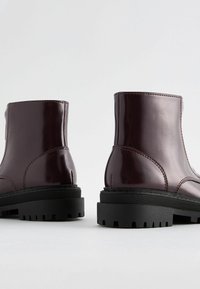 Stivali alla caviglia in pelle verniciata bordeaux con suola in gomma nera spessa. Cuciture evidenti e design elegante con punta arrotondata.
