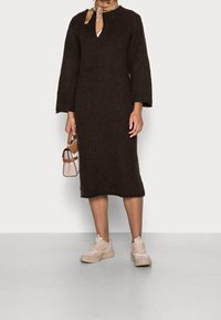 Braun gestricktes Kleid mit tiefem V-Ausschnitt und langen Ärmeln, kombiniert mit beigen Sneakers und einer kleinen, hellen Handtasche.