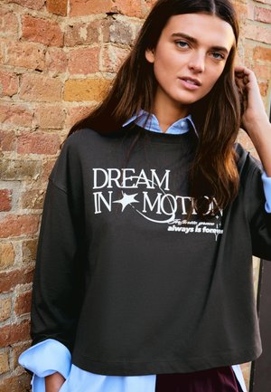 Fiatal nő hosszú barna hajjal áll egy téglafal előtt, fekete pulóvert visel, amelyen a "DREAM IN MOTION" felirat olvasható, világoskék ing alatt.