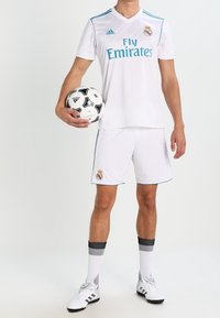 Valkoinen Adidas-jalkapalloharjoituspuku, jossa on sinisiä yksityiskohtia. Varustettu "Fly Emirates" -logolla, seuran merkillä, ja mustavalkoinen jalkapallo pidetään toisessa kädessä.