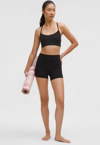 Fekete sportmelltartó és magas derekú short, mindkettő testhez simuló. A modellen rózsaszín és fehér csíkos jógászőnyeg van, mezítláb, semleges háttér előtt.