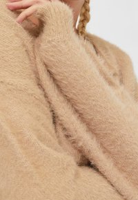 Beiger, flauschiger Strickpullover mit lockerem Schnitt, langen Ärmeln und gerippten Bündchen für zusätzliche Textur. Weiches Material mit einem warmen Erscheinungsbild.