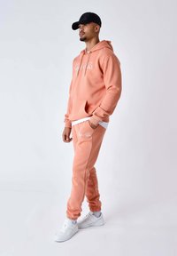 Sudadera con capucha color coral y pantalones deportivos a juego en tela suave, con bolsillo frontal y logo. Combinados con zapatillas blancas y una gorra negra.