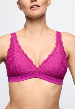 Vrouw die een felroze kanten bralette met bloemmotieven draagt, waarbij schouders en bovenborst tegen een effen achtergrond zichtbaar zijn.