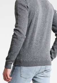 Grauer Strickpullover mit einem strukturierten Muster an den Ärmeln, gerippten Bündchen und Saum, kombiniert mit hellblauen Jeans.