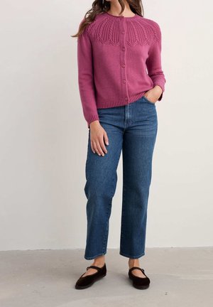 Vrouw draagt een roze gebreid vest met knopen en een getextureerde pasvorm, blauwe jeans met rechte pijpen, en zwarte Mary Jane schoenen, staand met hand in de zak.