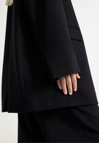 Blazer nero su misura con una texture liscia, caratterizzato da una tasca e una silhouette leggermente più lunga. Mano posata delicatamente sulla cucitura laterale.