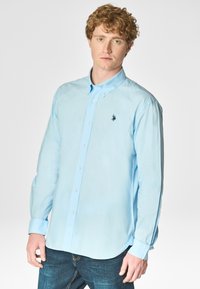 Camicia azzurra chiara con colletto, maniche lunghe e un piccolo logo scuro sul lato sinistro del petto. Tessuto di cotone con una trama liscia.
