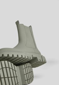 Chubby béžové kotníkové boty s poutky a silnými, vroubkovanými gumovými podrážkami vystavené na jednoduchém světlém šedém pozadí.