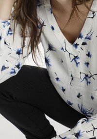 Blouse blanche avec motif floral bleu et accents noirs, présentant un col en V et des détails de boutons sur les manches. Portée avec un pantalon noir.