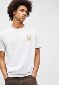 Λευκό βαμβακερό t-shirt με εκτύπωση σχεδίου "Notting Hill" και πολύχρωρα γραφικά στην αριστερή πλευρά του στήθους. Κοντά μανίκια, κλασική εφαρμογή.