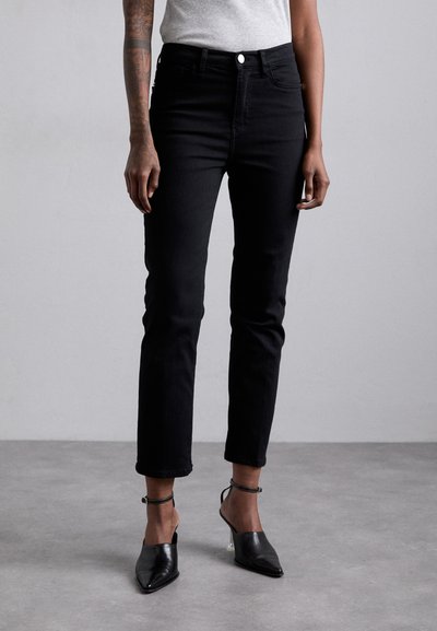 Schwarze Denim-Jeans mit einer körperbetonten Silhouette, hoher Taille, Fünf-Taschen-Design und dezenten Nähten. Kombiniert mit schwarzen Absatzmules.