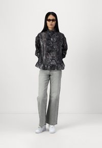 Blusa nera con stampa serpente e scollatura a volant, abbinata a jeans a gamba larga grigi chiari e sneakers bianche. La modella indossa occhiali da sole scuri.