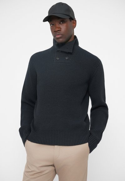 Tummansininen neulottu villapaita, jossa on korkea kaulus, ribatoitu pinta ja kaksi nappia kauluksessa. Yhdistetty vaaleisiin housuihin.