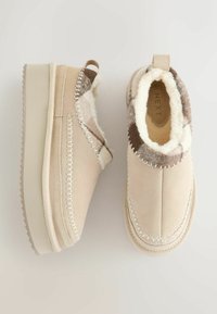 Beige suède instap schoenen met een gestructureerde bruin-witte bontaccent, voorzien van witte stiksels langs de zijkanten en een stevige rubberen zool.
