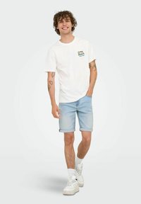 Bijela pamučna majica s grafikom "Long Beach Surf Club", u kombinaciji sa svijetloplavim trapericama i bijelim tenisicama. Ležeran kroj i rolane rubove.