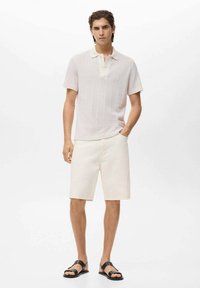 Lyse beige strikket polo skjorte med korte ærmer og knapkrave, parret med cremefarvede shorts og sorte sandaler, stående mod en hvid baggrund.