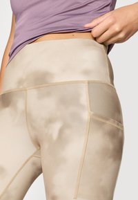 Μπεζ κολάν με ψηλή μέση και ομαλή υφή, με σχέδιο tie-dye και αντίθετες ραφές. Συνδυασμένο με ένα ανοιχτό μωβ τοπ.