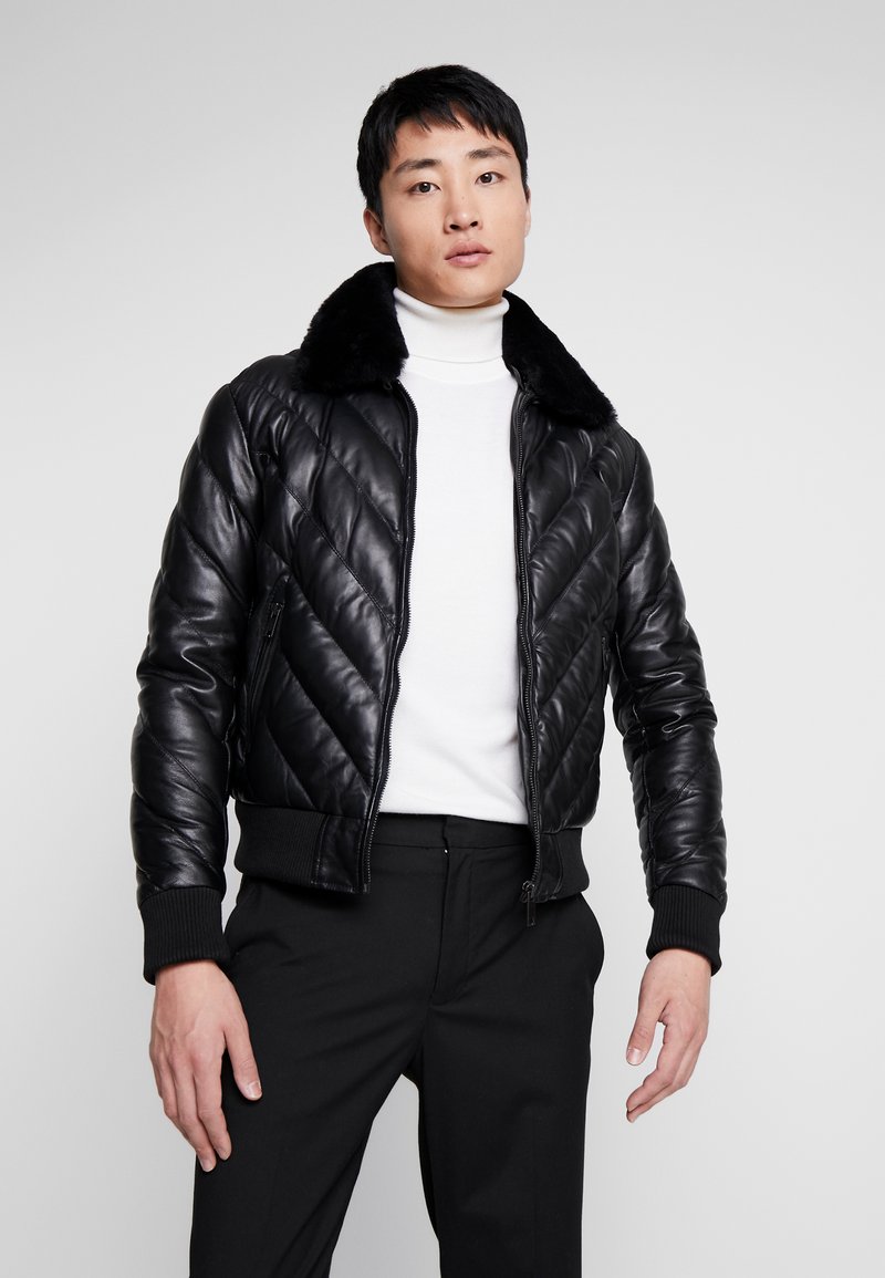 Zwarte leren bomberjack met een gequilt ontwerp, voorzien van een nepbont kraag en geribbelde manchetten. Model draagt een witte coltrui en donkere broek.