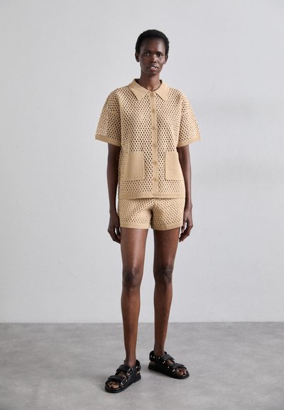 Beige crochet two-piece outfit; short-sleeved button-up top with pockets, matching shorts. Model wears black sandals, standing on a gray surface.