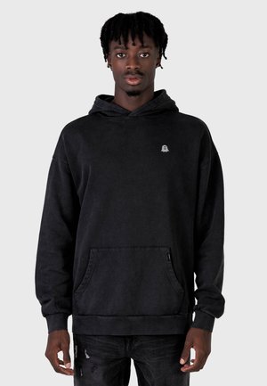 Giovane uomo dalla pelle scura e capelli intrecciati che indossa una felpa nera con cappuccio con un piccolo logo a forma di fantasma e jeans neri strappati.