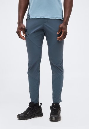 Kék sportnadrágot és fekete terepfutó cipőt viselő férfi, fehér, egyszínű háttér előtt áll.