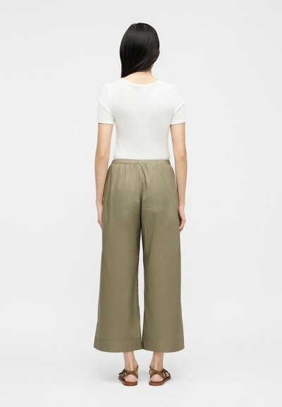 Ženska z dolgimi črnimi lasmi, oblečena v oprijeto belo majico s kratkimi rokavi, široke ohlapne olivno zelene hlače in rjave sandale, stoji obrnjena stran.