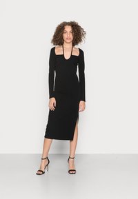 Vestido preto de manga longa com decote arredondado, silhueta ajustada, comprimento até as panturrilhas e uma fenda lateral. Usado com sapatos de salto pretos com tiras.