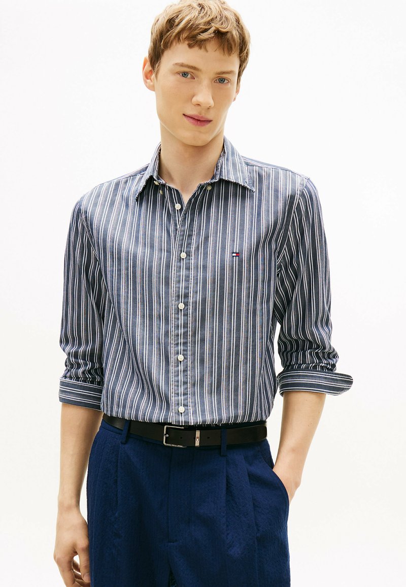 Jeune homme portant une chemise à manches longues à rayures verticales bleues et blanches avec les manches retroussées et un pantalon bleu foncé, debout avec une main dans la poche.