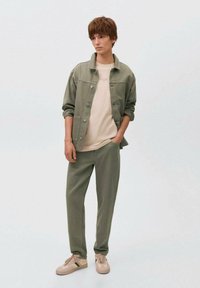 Olivno zelena denim jakna, bež majica s tekstom, usklađene zelene hlače i svijetle bež tenisice, sve u ležernom kroju.