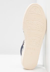 Semelle d’une sandale espadrille beige clair avec des motifs texturés et une marque, présentée sur un fond blanc et un coin de mur.