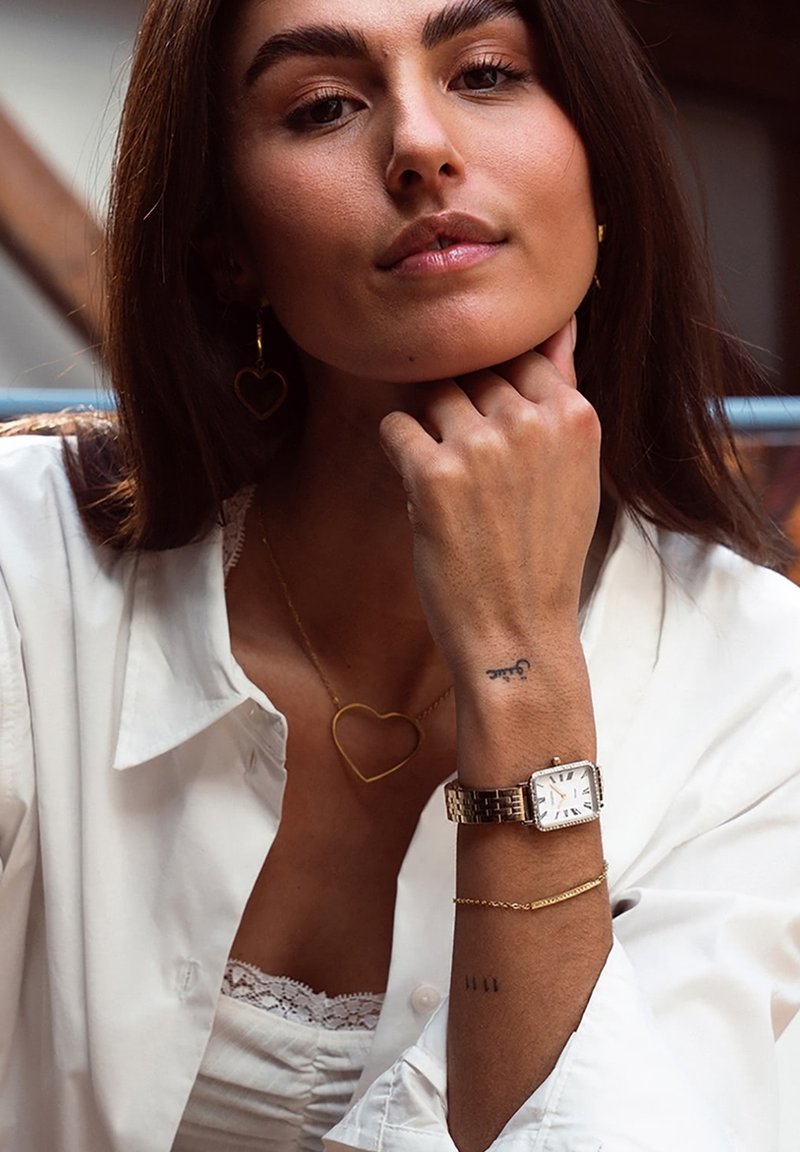 Montre rectangulaire en or avec un cadran blanc, chaînes superposées comprenant un pendentif en forme de cœur, et boucles d'oreilles. Présente une peau lisse et une chemise en tissu léger.