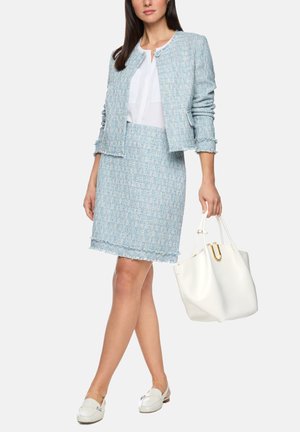 Frau trägt hellblauen Tweed-Rockanzug mit weißer Bluse, hält eine weiße Handtasche und trägt weiße Loafers, steht vor weißem Hintergrund.