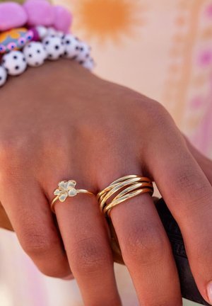 Mano con dos anillos de oro, uno en forma de flor y otro con varias bandas, y pulseras con cuentas blancas y moradas.