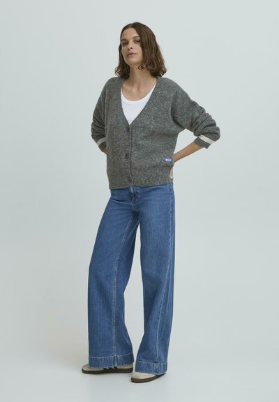 Sivý pletený cardigan s výstrihom do V, prednými gombíkmi a nášivkou loga, v kombinácii s modrými širokými nohavicami s vysokým pásom a béžovými topánkami.