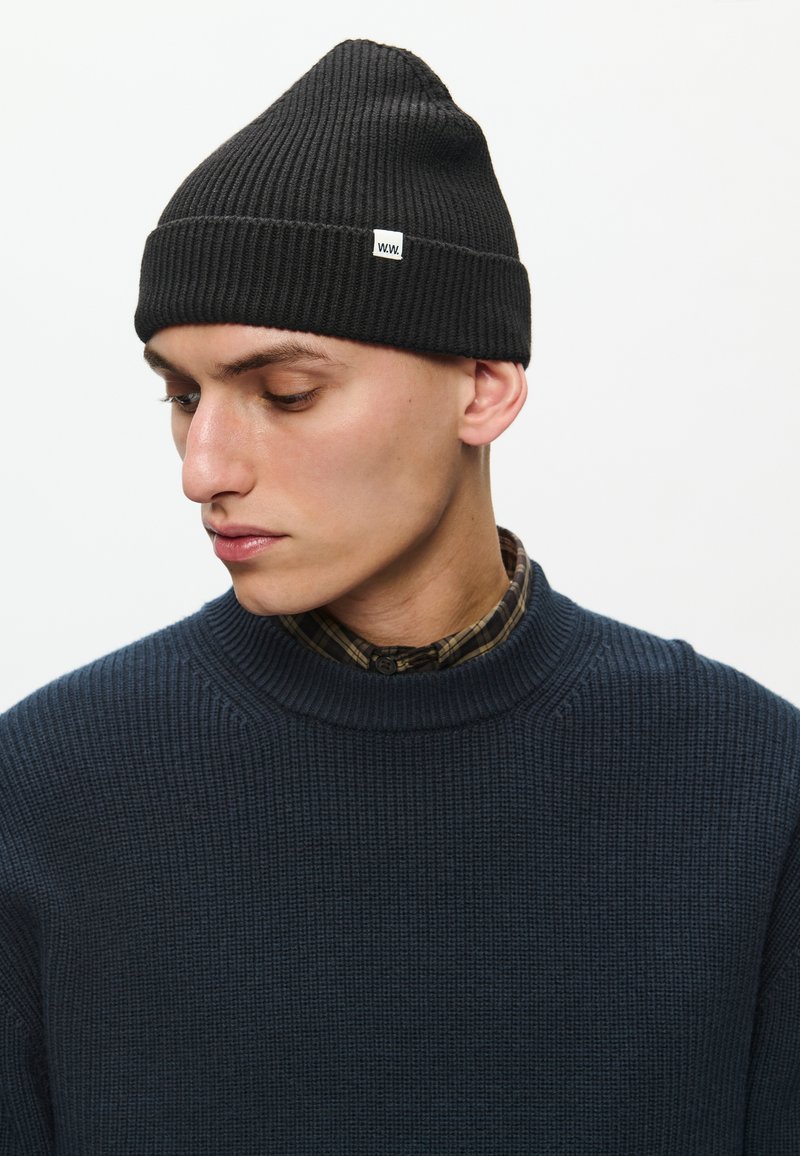 Black ribbed knit beanie with a pointed top, featuring a small white label. Worn with a textured navy sweater and a plaid shirt underneath.