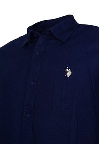 Námornícká modrá košeľa na gombíky s golierom a malým bielym vyšitým logom polového hráča na koni na ľavej hrudi.