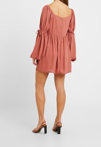 Malinové růžové šaty s nafouknutými rukávy a čtveratým výstřihem na zádech, s vypasovaným pasem a vrstveným designem. V páru s béžovými otevřenými lodičkami.