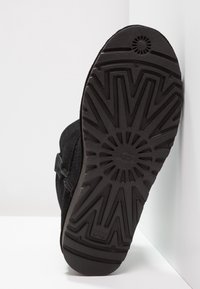 Czarna podeszwa buta UGG z promienistym wzorem bieżnika, ustawiona pod kątem względem białej ściany i podłogi.