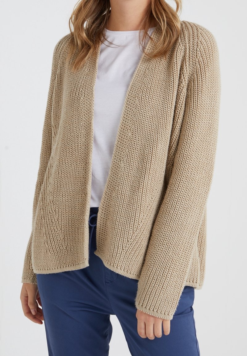 Beige open-front vest met een gestructureerd breipatroon, verlaagde schouders en geribbelde manchetten, gecombineerd met een wit T-shirt en een blauwe broek.