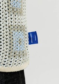Gros plan de l'ourlet d'un pull beige à maille ajourée avec étiquette bleue Jack & Jones Originals au-dessus d'un jean en denim noir.
