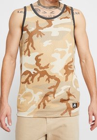 Débardeur en mesh beige avec un motif camouflage dans des tons de beige et de marron. Il présente un col noir et un petit logo noir au bas.