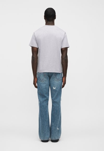 Sivá tričko s krátkym rukávom v kombinácii s poškodenými svetlomodrými rozšírenými džínsami, ktoré majú mierne skrátený strih a dve zadné vrecká.