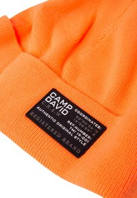 Orange knitted beanie with ribbed texture and a black rectangular tag featuring white text, including brand name and coordinates.