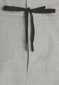 Grijze stof met een subtiele textuur, voorzien van een zwarte trekkoord met metalen aglets en een kleine metalen accent bij de tailleband.