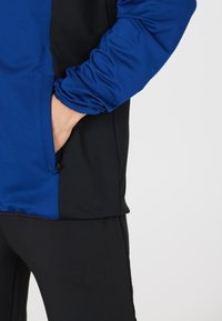 Blå og sort atletisk jakke med en glat tekstur, med en sidelomme og elastiske ærmer, delvist stikkende ind i sorte bukser.