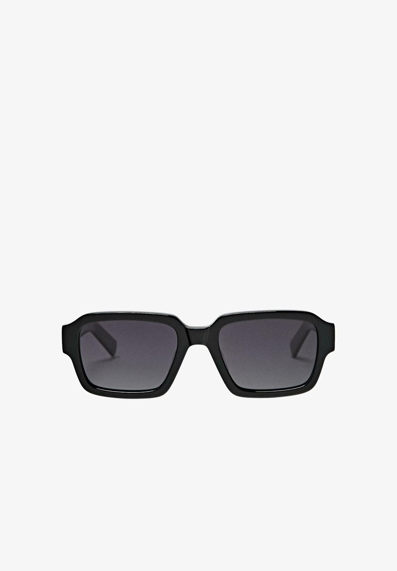 Lunettes de soleil rectangulaires noires avec des montures épaisses et des verres teintés foncés sur fond blanc.